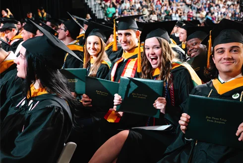 A diverse group of graduates in caps and gowns joyfully holding their diplomas during a commencement ceremony.