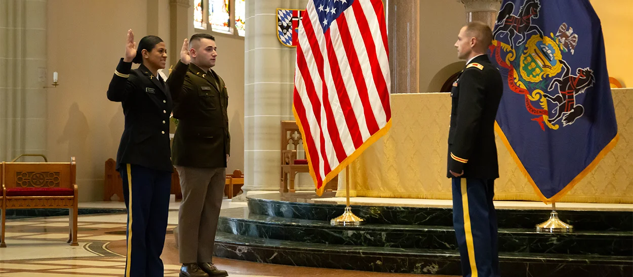 Three individuals in military uniforms participate in a ceremony inside a church-like setting, with flags displayed behind them. 