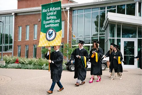 Graduates from the Aler G. McKenna School of Business, Economics, and Government march in a procession holding a banner.