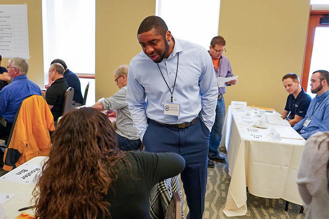 A KCOE workshop scene featuring a man in a blue shirt engaging with a participant, surrounded by attendees at tables.