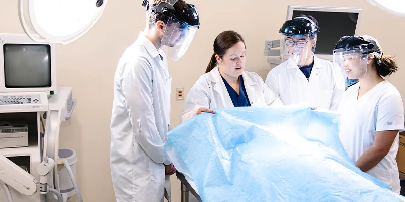 Medical students collaborating in a surgical setting, wearing protective gear and preparing a sterile surgical drape.