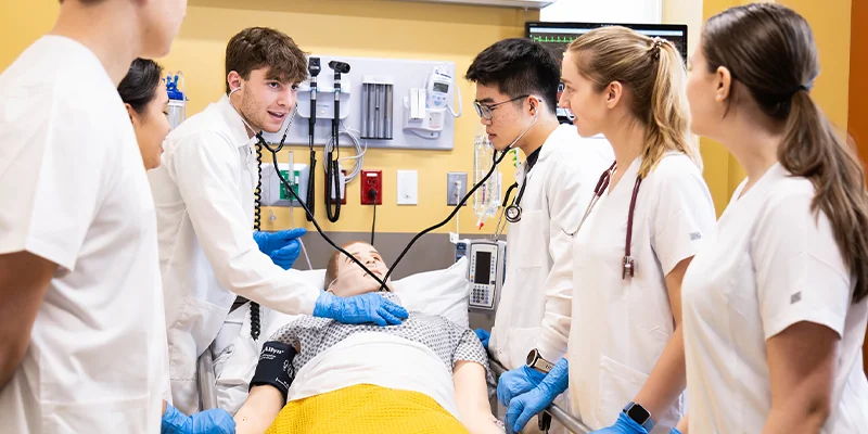 Medical students in scrubs collaborating around a simulated patient in a clinical setting.