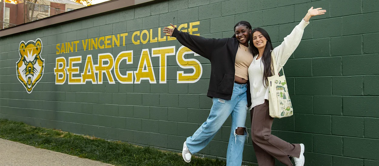 Two students pose joyfully in front of a mural featuring the Saint Vincent College Bearcats logo.