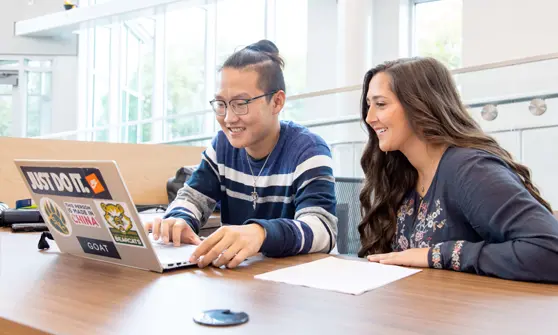 two students sitting at a table looking at a laptop screen