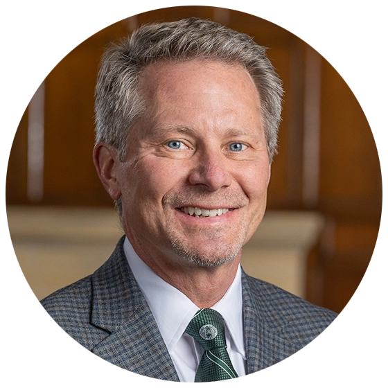 Portrait of Kevin Guskiewicz smiling in a checked suit jacket and tie, set against a wooden background.