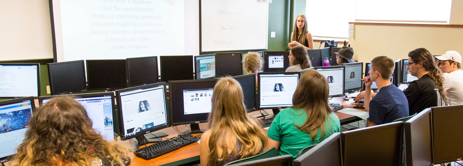 A female teacher stands at the front of a classroom and speaks to teenage students who each have a computer