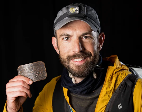 A smiling man in a yellow jacket displays a vintage belt buckle, showcasing his achievement in an outdoor adventure.