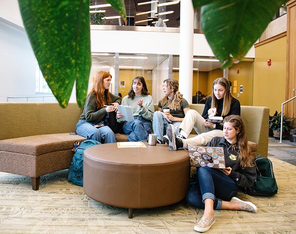 Students gathered around a couch in a lobby area looking at laptop screens.