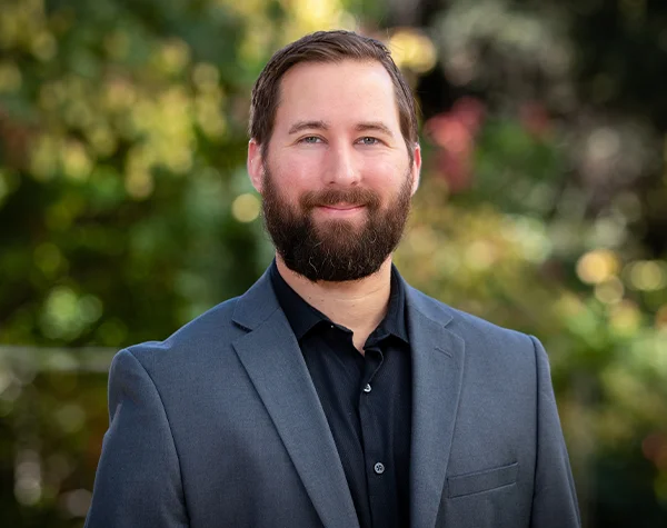 Professional headshot of a man with a beard, wearing a gray suit and black shirt, posing outdoors with greenery in the background.