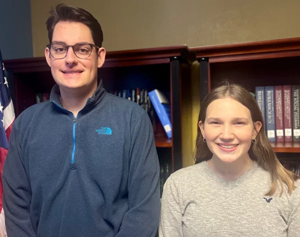 Two students standing side by side, smiling in front of a bookshelf, with an American flag visible to the left.