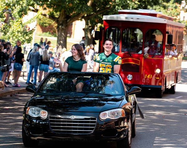 Photo of homecoming students riding in a car with trolly behind them and students lookin on and taking photos