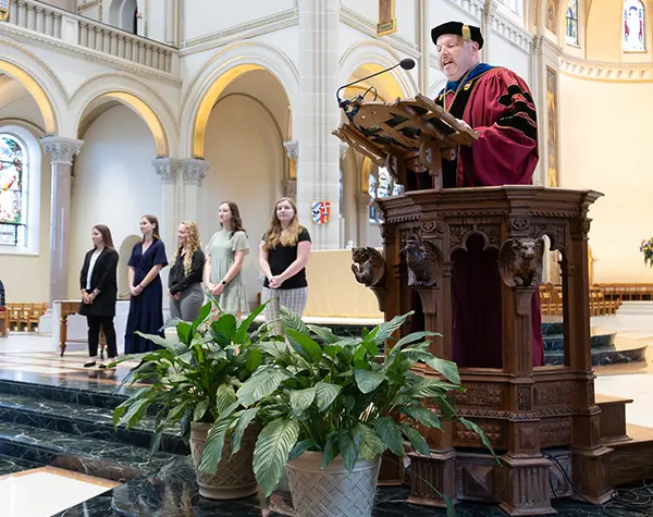 Rev. Paul Taylor President Saint Vincent College speaking at the Honors Convocation in the Basilica with a group of studnts standing to his right.