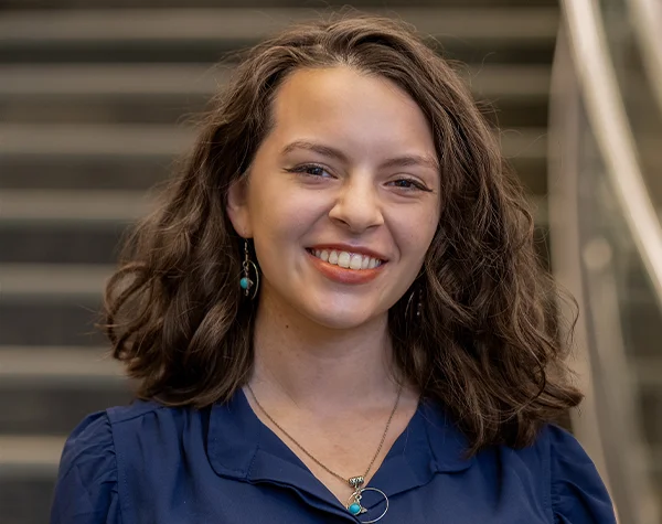 Smiling woman with curly hair, wearing a blue blouse and turquoise earrings, stands in front of a staircase.