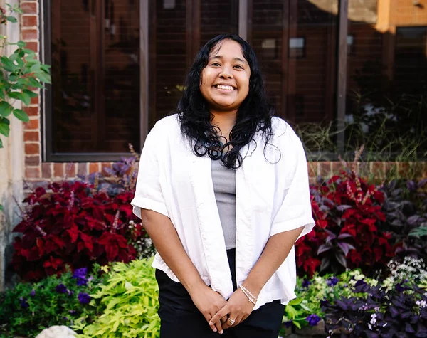 A smiling young woman stands in front of a vibrant display of plants and flowers, wearing a white shirt and black pants.