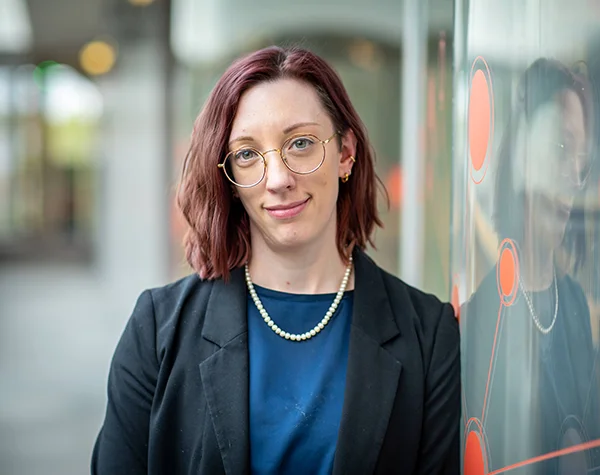 Professional headshot of a woman with short, auburn hair and glasses, dressed in a black blazer and pearl necklace, standing against a modern glass backdrop.