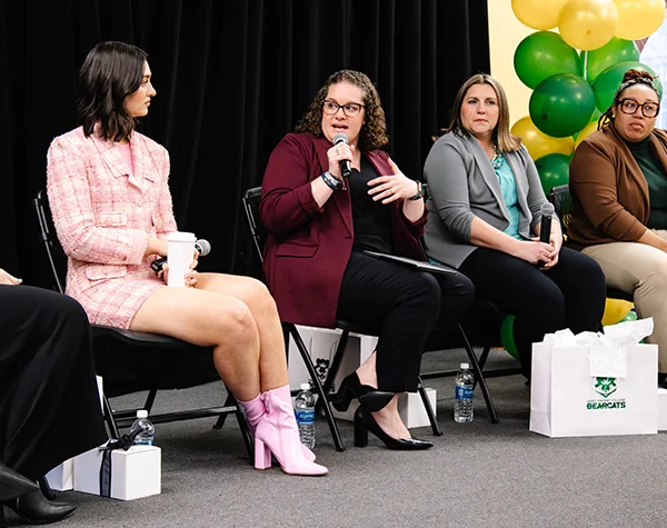 A panel discussion featuring four women, with one speaker in a maroon blazer and glasses addressing the audience.