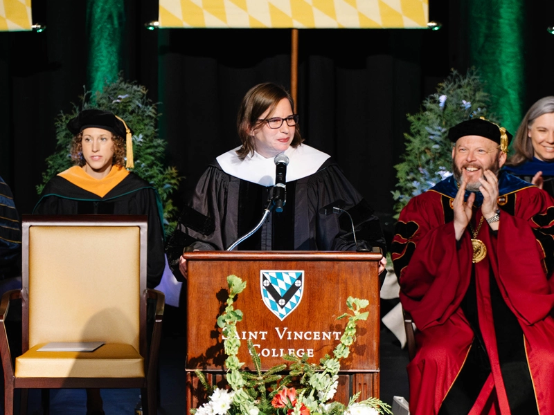 A formal graduation ceremony featuring speakers and faculty members in academic regalia, positioned at a podium with banners representing various schools.