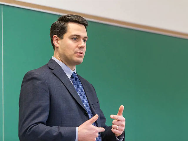 Dr. Jerome Foss gestures while speaking in front of a green chalkboard.