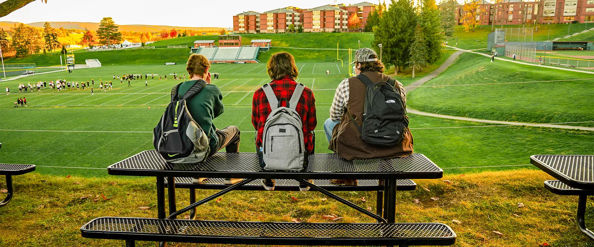 Three students sitting on a bench, overlooking a grassy field and sports practice area during autumn. The scene captures a relaxed outdoor atmosphere on campus.