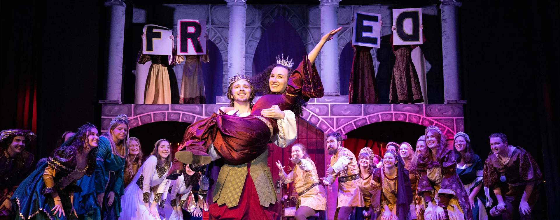 A lively theater scene featuring actors in elaborate costumes during a performance, with one actor lifting another in front of a colorful backdrop .