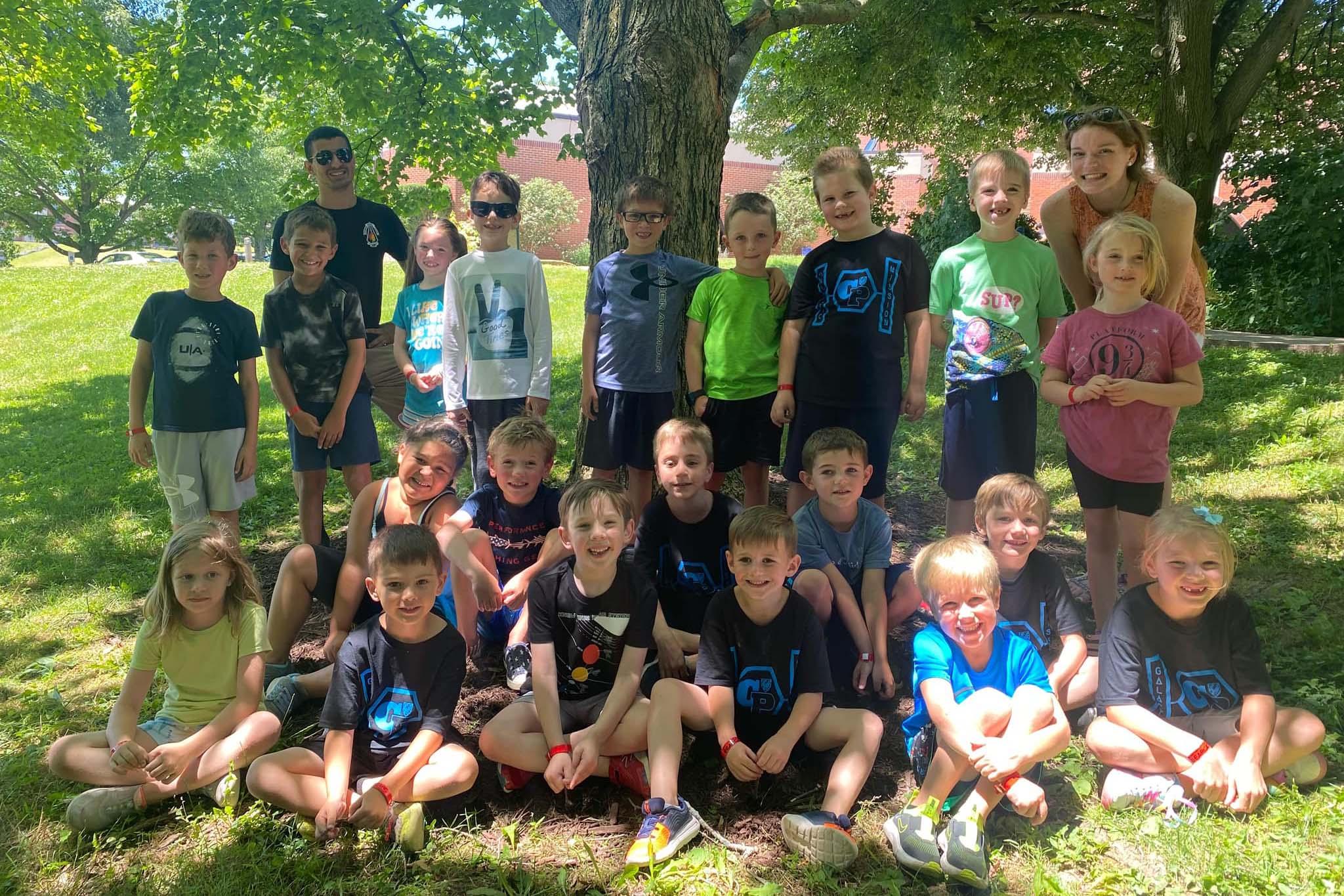 Group photo of children and adults smiling together in a grassy area under a tree during a camp or outdoor activity.