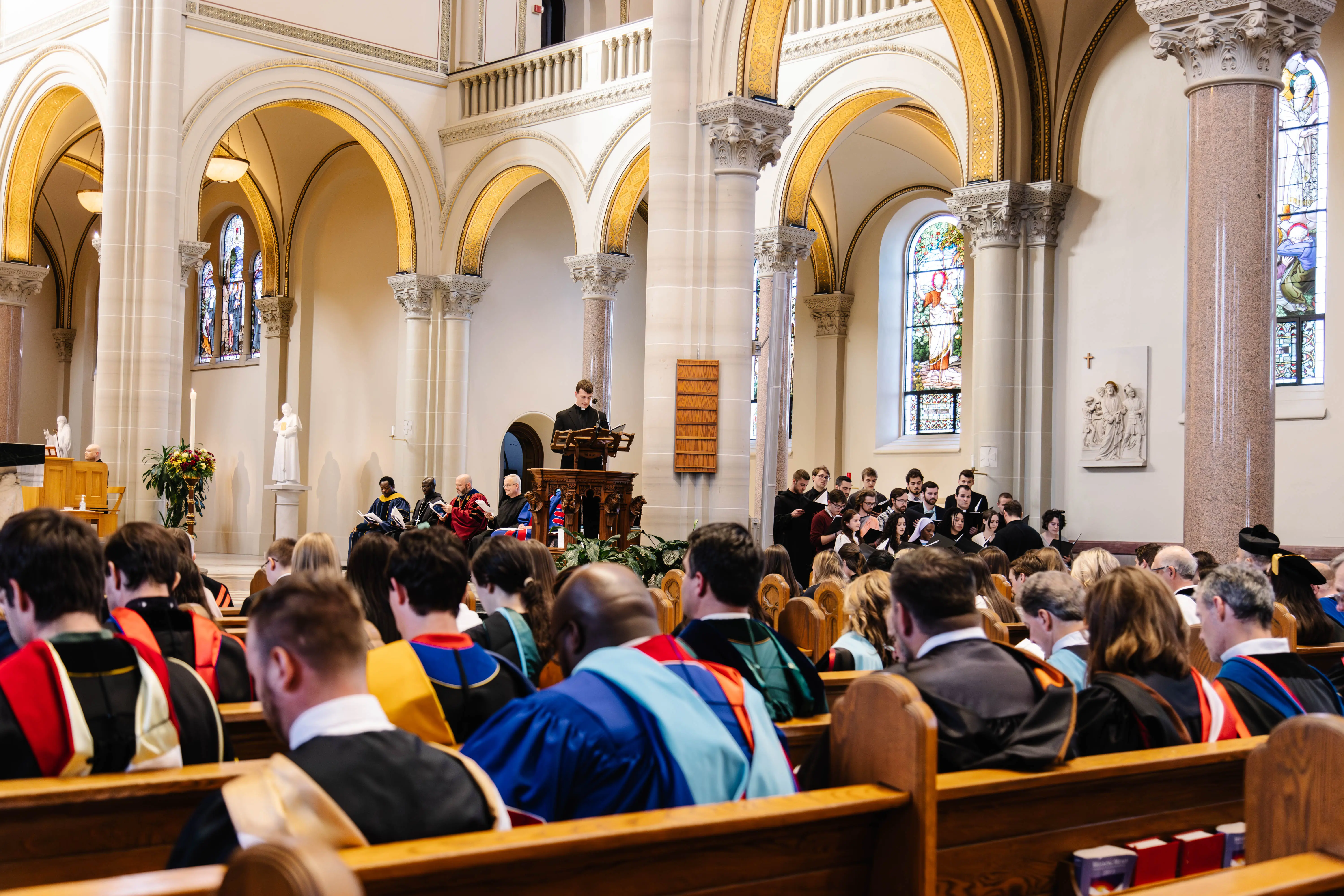 A formal gathering in a church setting, featuring faculty members in academic regalia seated in pews, with a speaker at a podium and a choir in the background.