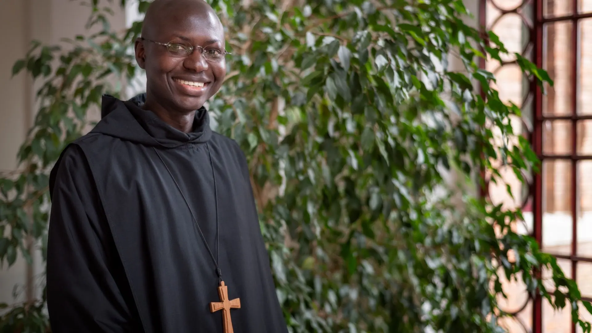 Portrait of a smiling monk in black robes, wearing a cross necklace, standing in front of lush greenery.