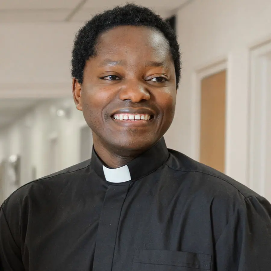 A smiling priest in a black clerical shirt and white collar, standing in a hallway.