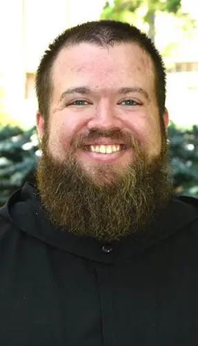 A smiling man with a full beard, wearing a black hooded garment, stands outdoors against a blurred green background.