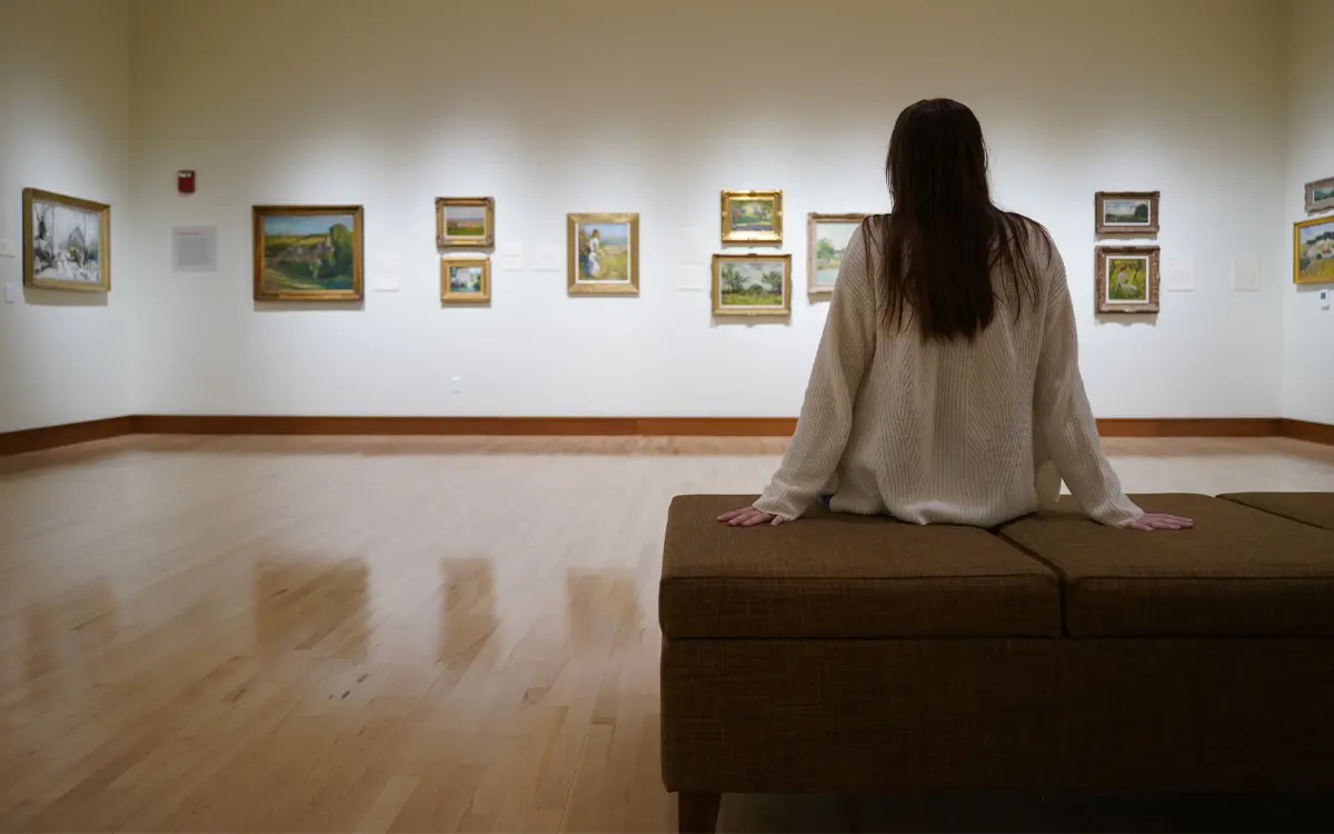 A visitor sits on a bench in an art gallery, admiring a collection of framed paintings on the wall.