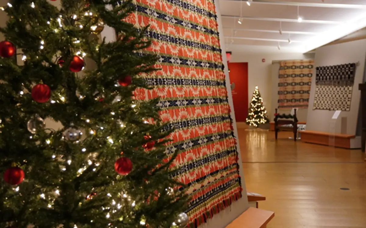 Interior view of a museum exhibition featuring colorful woven tapestries and decorated Christmas trees.