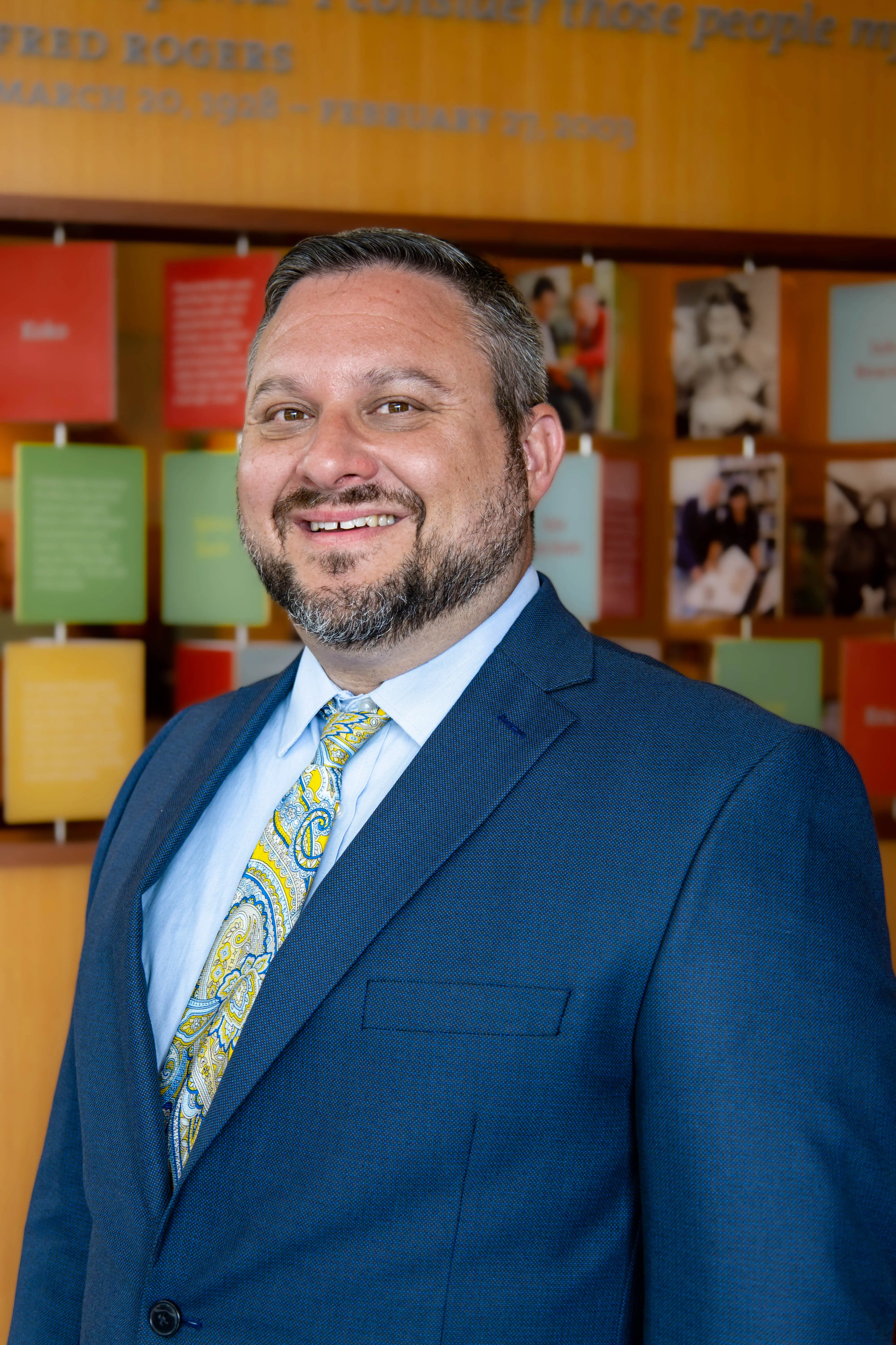 A professional portrait of a man in a suit and tie, smiling confidently against a colorful backdrop of framed quotes and photographs.