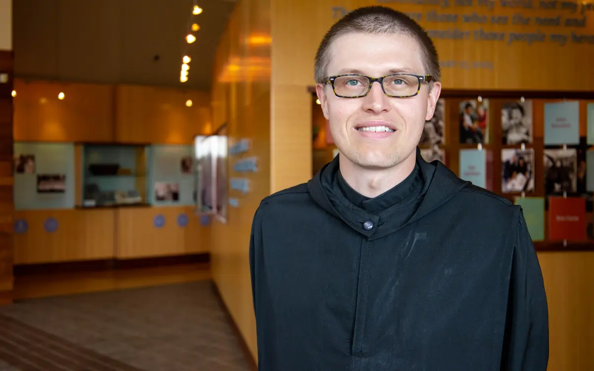 A smiling man with glasses stands in a well-lit lobby, featuring a wooden interior and photos on the wall.