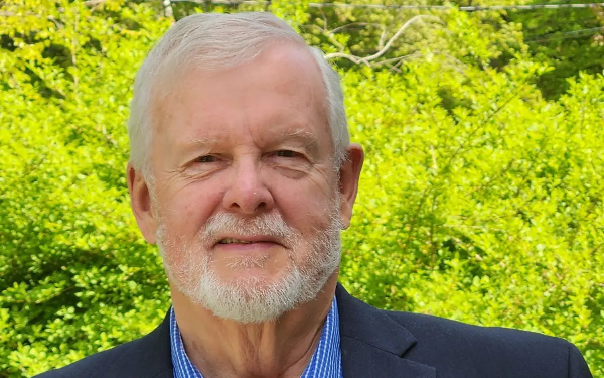 A senior man with a gray beard and light hair, wearing a blue shirt and dark blazer, stands in front of lush green foliage.