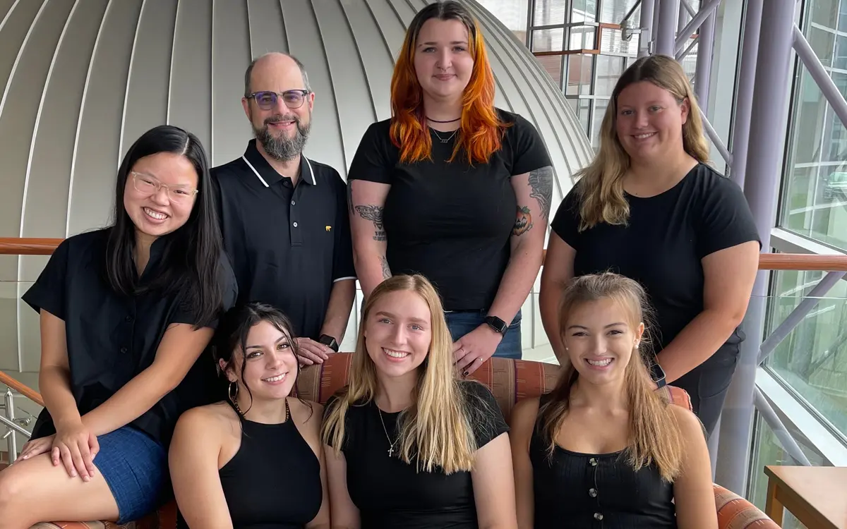 A group photo of seven people, all wearing black tops, posing together in a modern indoor setting.