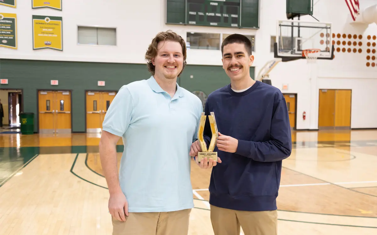 Two young men stand in a gymnasium, holding a trophy and smiling, celebrating an achievement.