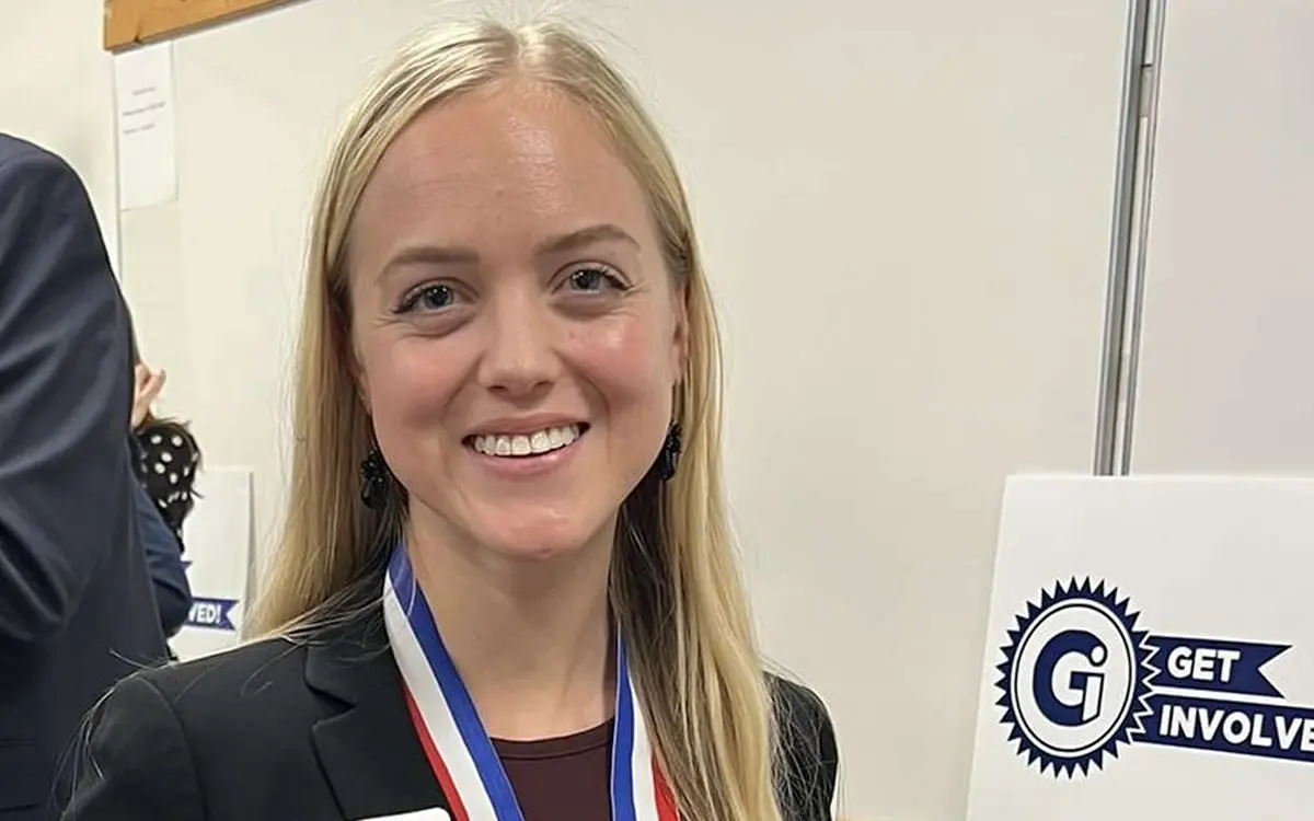 A young woman in a professional outfit smiles while wearing a medal and standing next to a "Get Involved" sign.