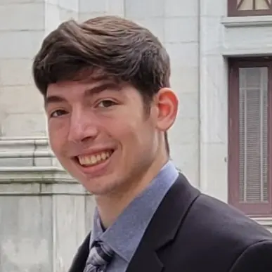 A young man with short brown hair smiles confidently while wearing a suit and tie, standing in front of a historic building.