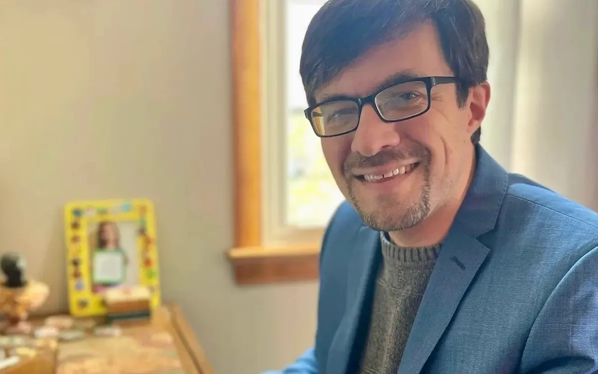 A smiling man in a blue blazer and glasses sitting at a desk, with a framed photo and decorative items in the background.