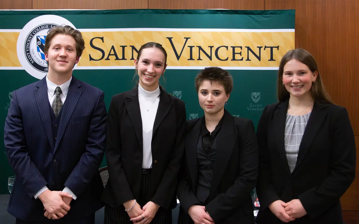 A group of four students dressed in professional attire posing in front of a Saint Vincent College backdrop.