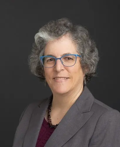 Professional portrait of a woman with curly gray hair and blue glasses, wearing a gray blazer, smiling against a dark background.