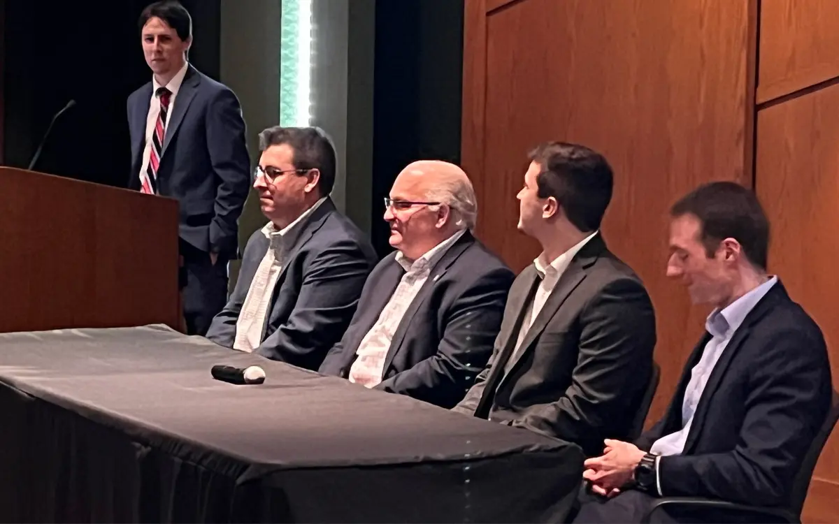 A panel of speakers seated at a table during a conference, with one speaker standing at the podium.