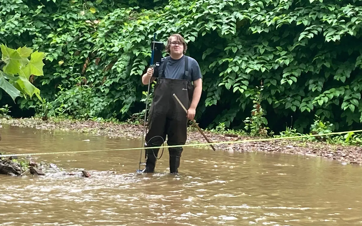 A person wearing waders stands in a flooded area, holding a surveying instrument and a stick, with dense greenery in the background.