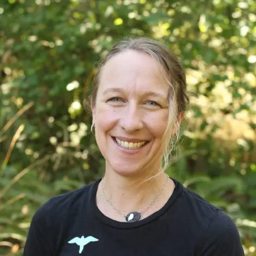 A smiling woman with long hair standing against a green blurred background, wearing a black shirt with a logo.