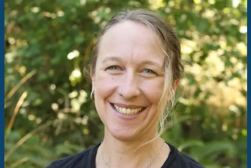 A smiling woman with long hair standing against a green blurred background, wearing a black shirt with a logo.