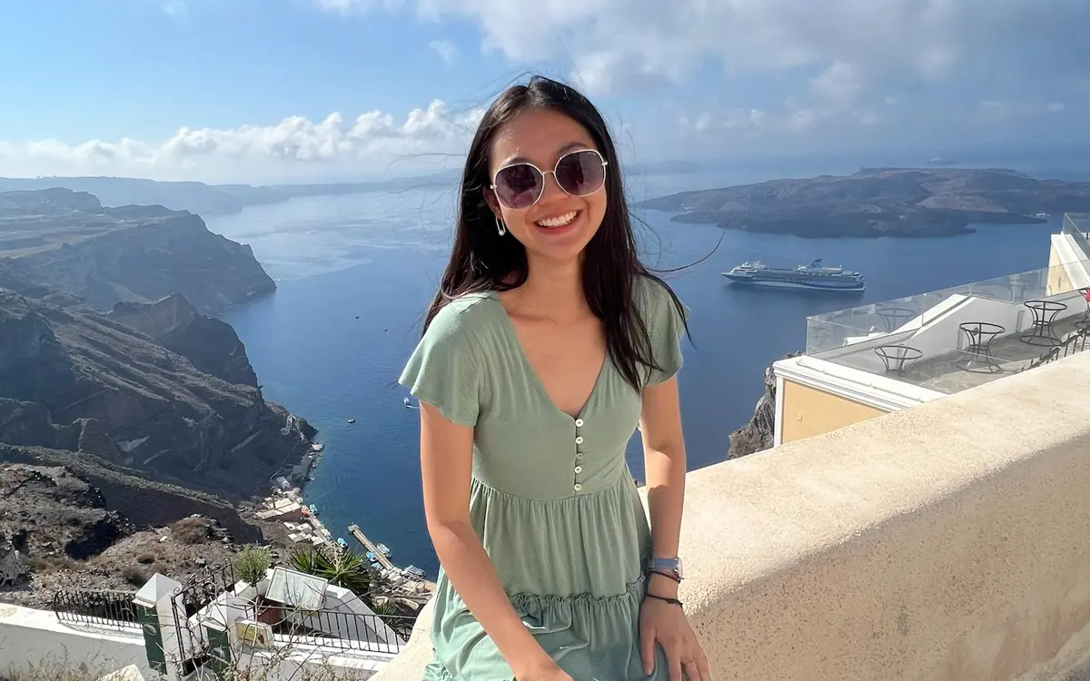 A young woman wearing sunglasses smiles while sitting on a ledge with a scenic view of the sea and cliffs in Santorini, Greece.