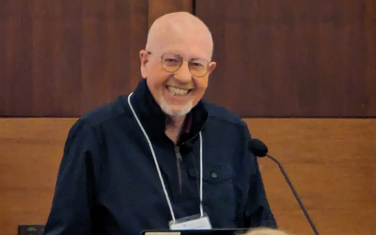 A smiling man with glasses and a bald head stands at a podium, speaking at an event.