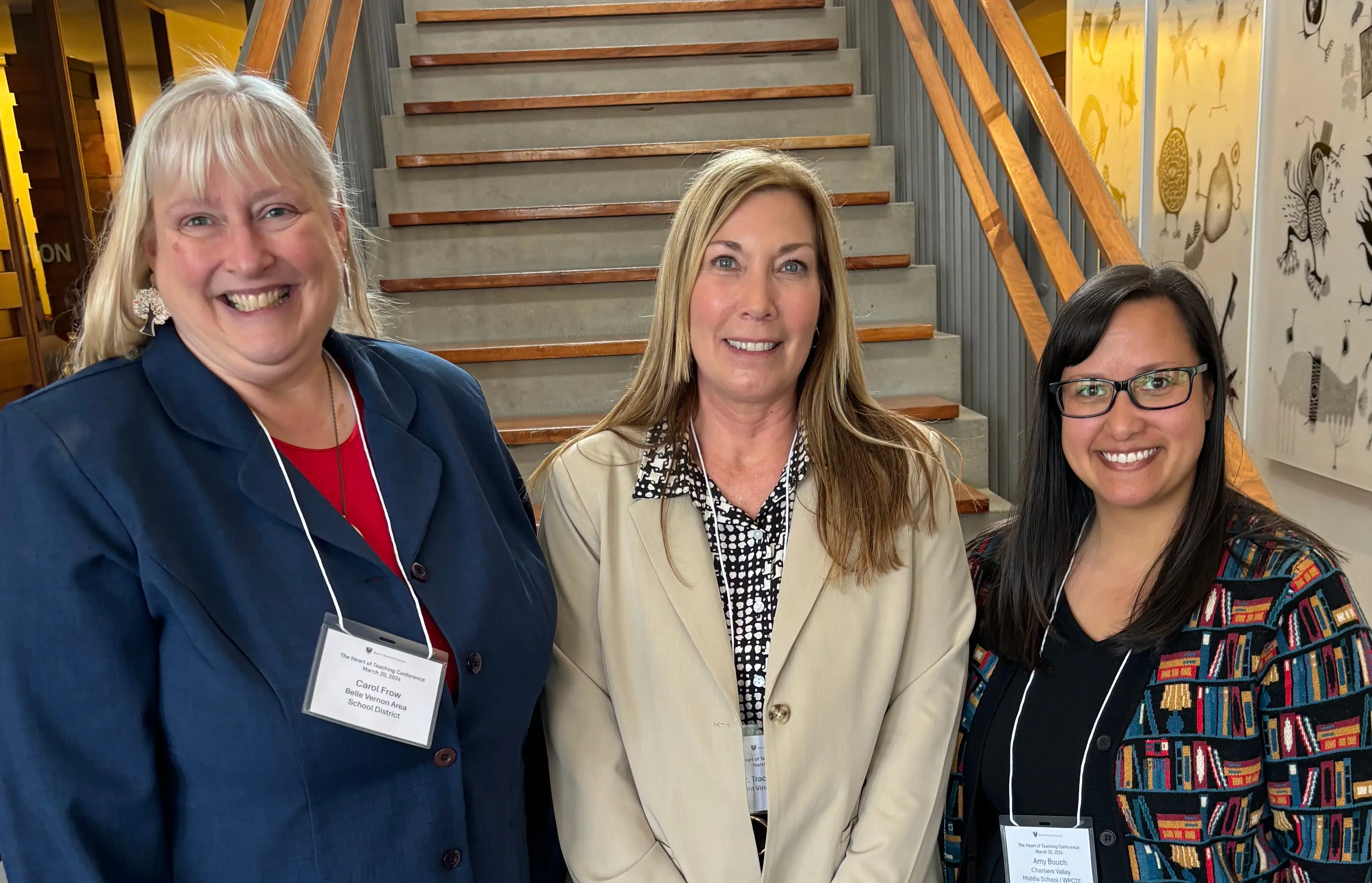 Three professional women smiling together at a conference, standing in front of a staircase.