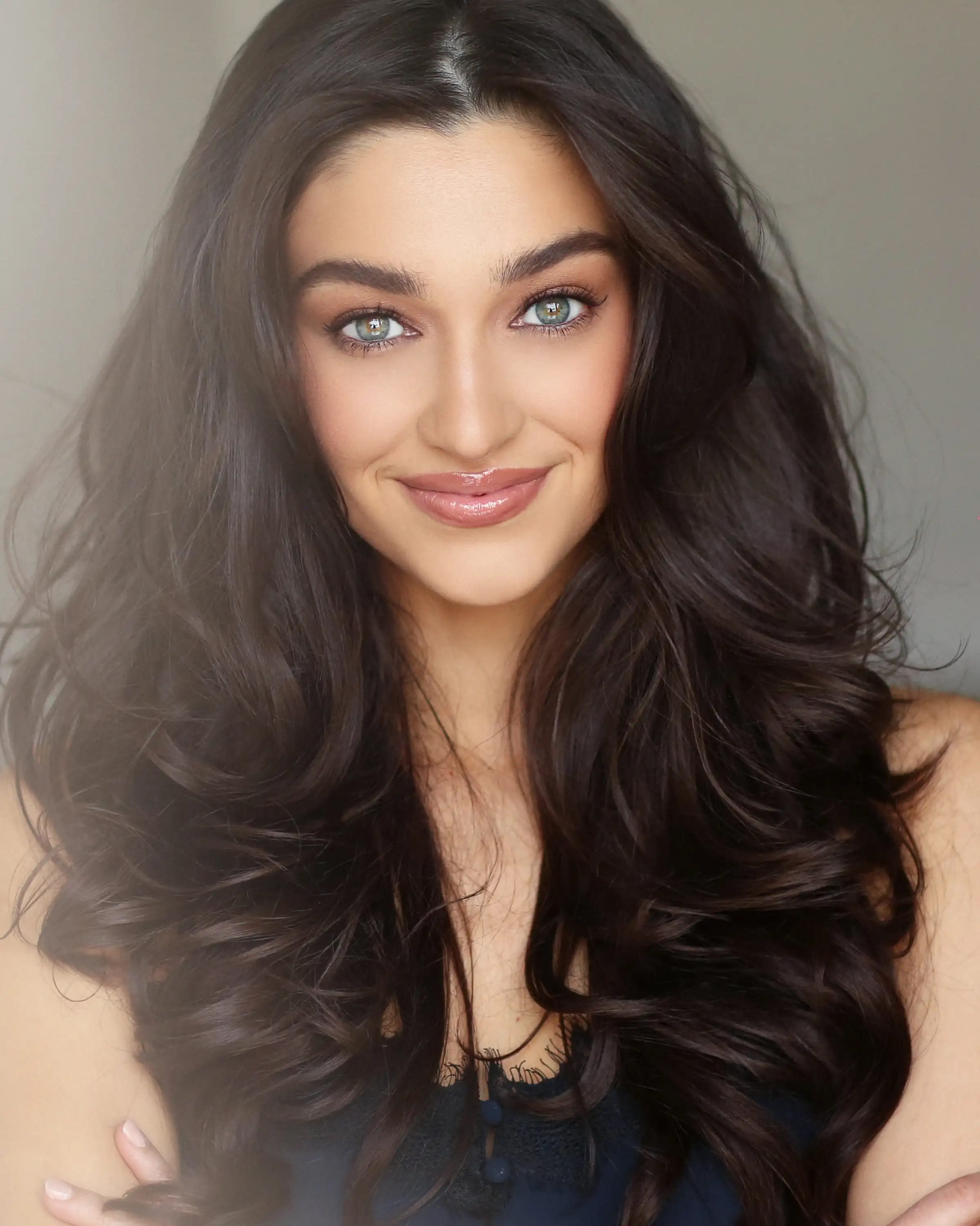 A woman with long, voluminous brown hair and bright blue-green eyes smiles warmly.