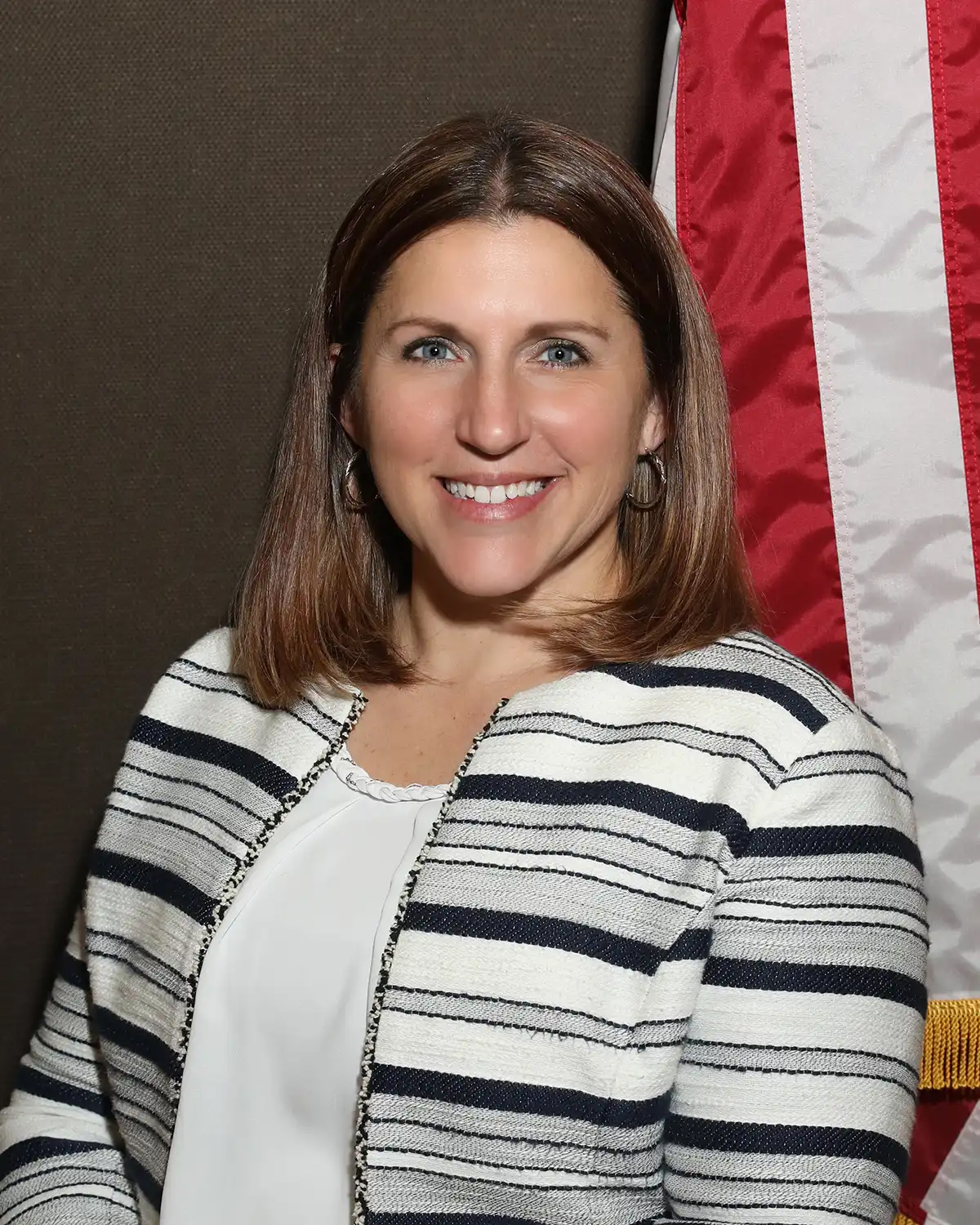 Professional portrait of a woman with shoulder-length hair, wearing a striped blazer, smiling in front of an American flag backdrop.